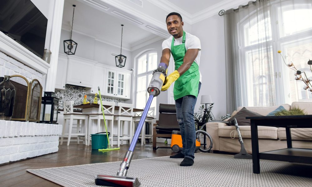 African man removing dust from soft carpet with modern vacuum cleaner. Male cleaner wearing green apron and yellow rubber gloves. Working process at bright room.