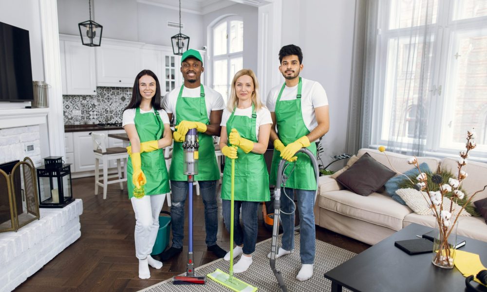 Group of four multiracial janitors in green aprons and yellow rubber gloves standing at modern apartment with cleaning equipment. Concept of professional service.