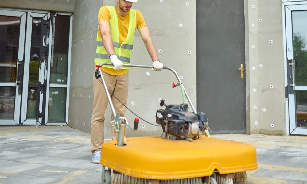 Cleaning. Man in protective helmet and gloves operating special equipment with brush cleaning construction site in new housing complex outdoors